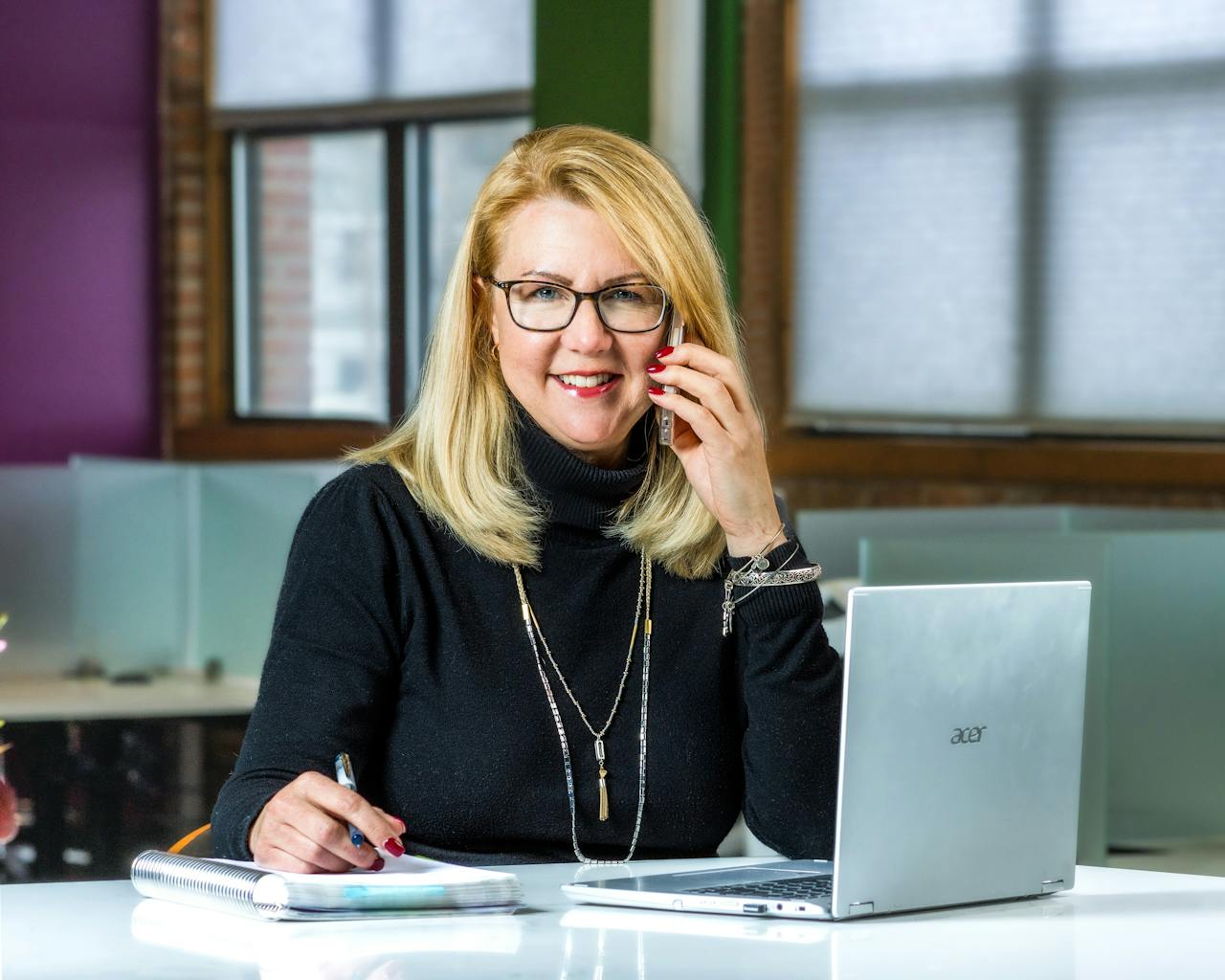 Smiling businesswoman on phone call, working on laptop in sleek office setting.